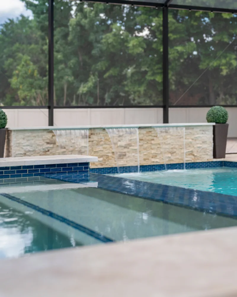 Custom pool with a stacked stone water wall and ivory travertine decking built by Pool Perfection in Odessa